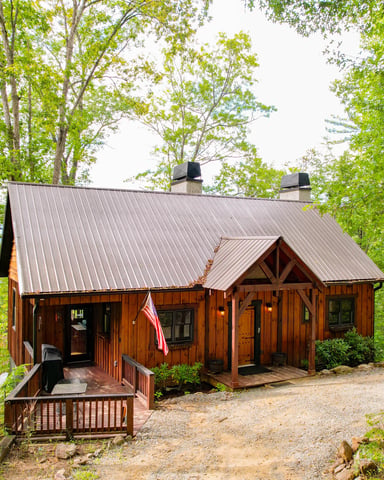 Green and white cottage with wraparound porch, two dormers, and stone chimney surrounded by dense forest and manicured lawn.