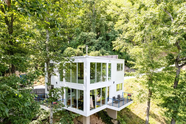 Modern white cubic house with large windows nestled among dense green trees, featuring a deck with outdoor furniture.
